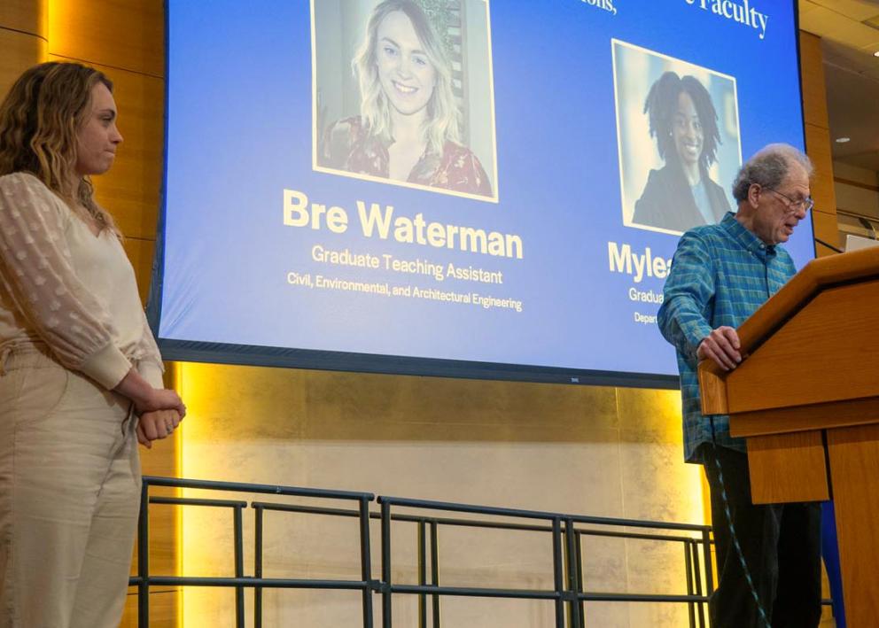Bre Waterman stands on stage while Dan Bernstein speaks at a poduim.